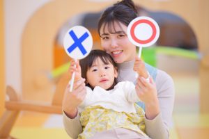◯・✕の札を持つ保育士とこども